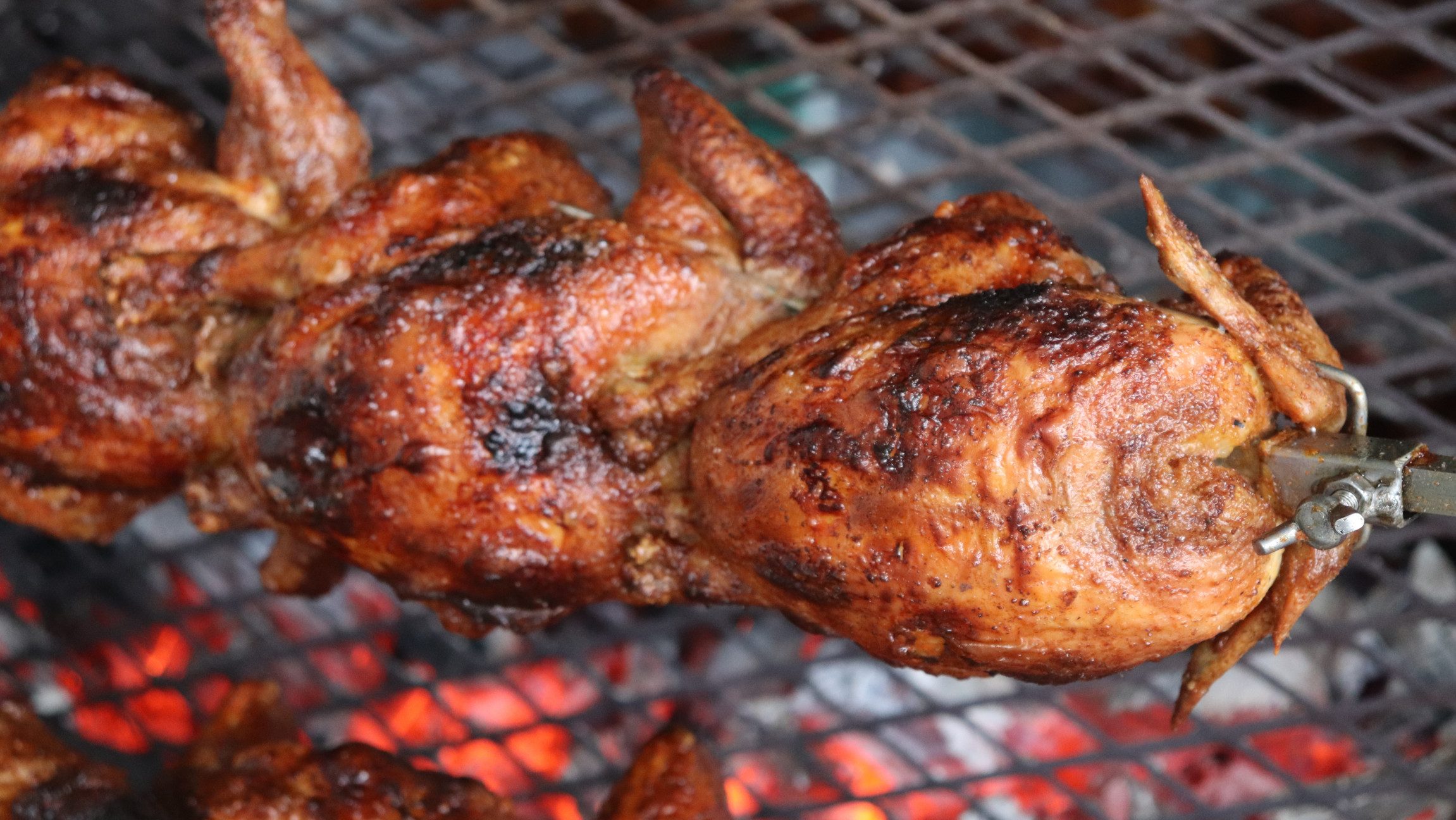 Blick auf einen Spieß mit Grillhendln, der über dem Feuer brät.