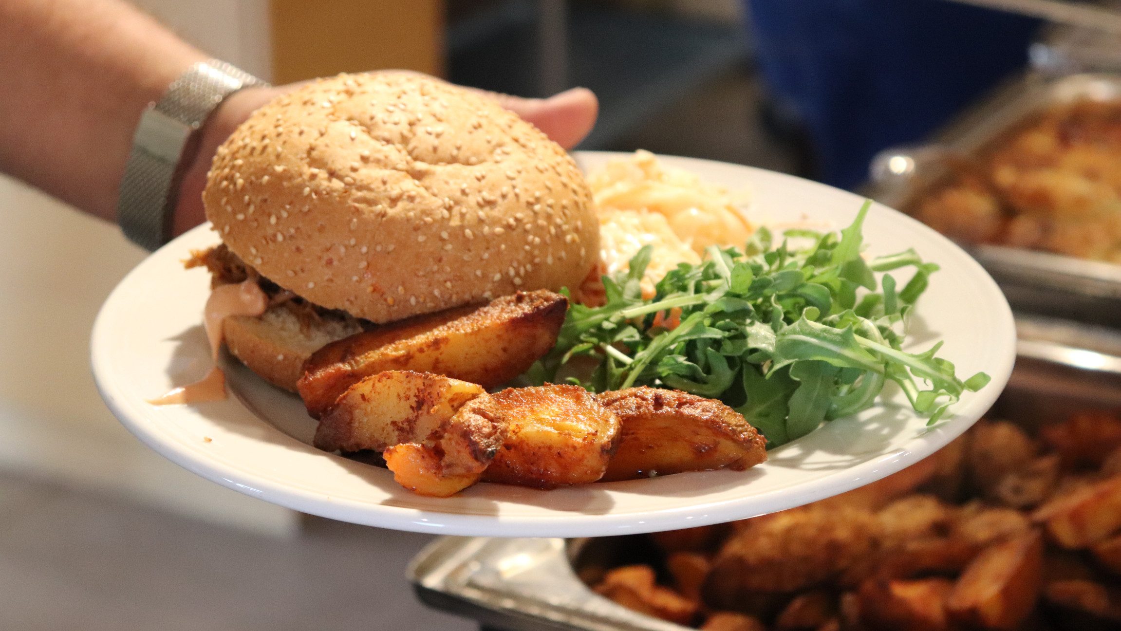 Blick auf ein Buffet vor dem jemand ein Teller mit Burger und Wedges in der Hand hält.