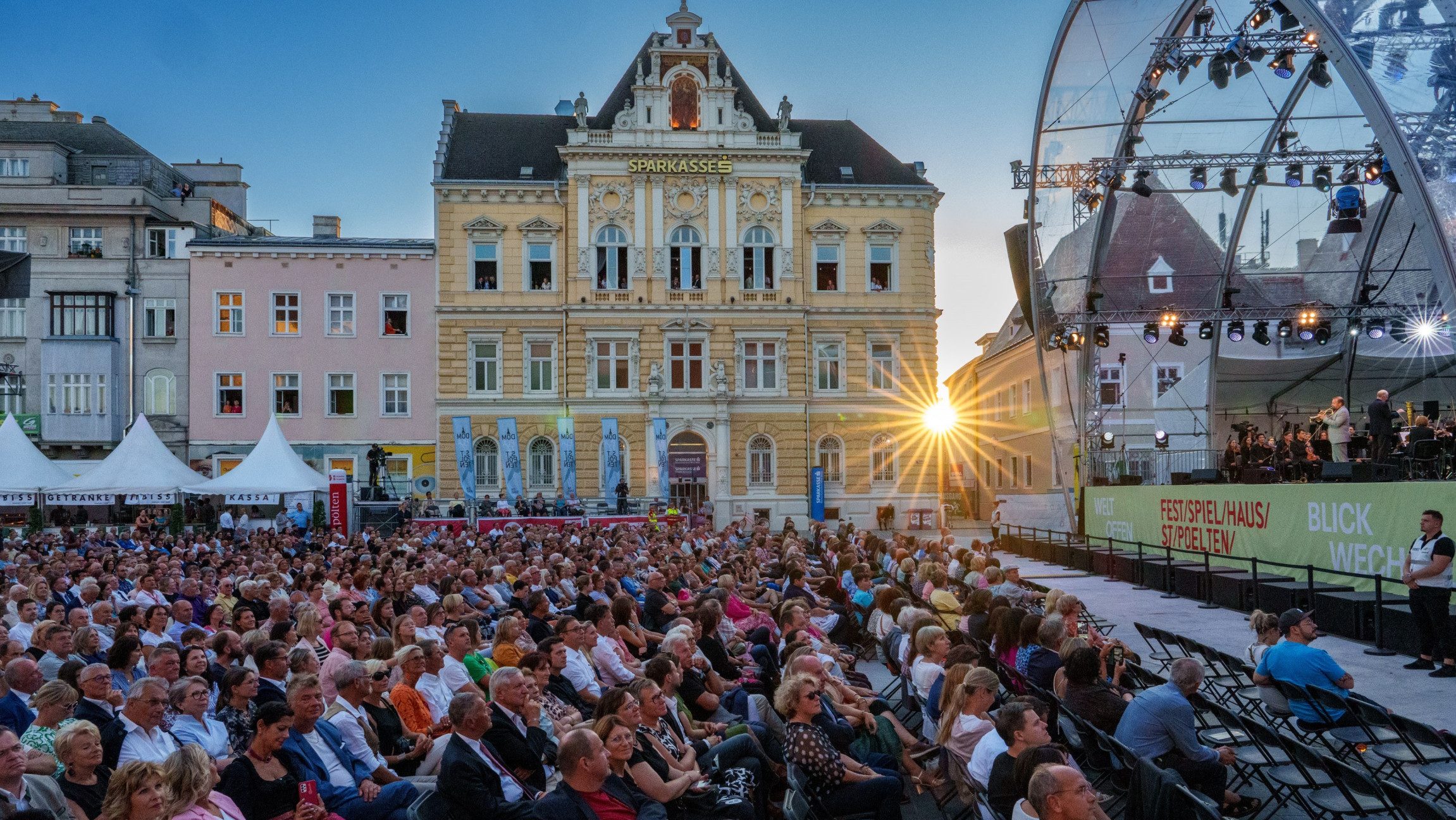 Blick auf ein Publikum, das vor einer Bühne sitzt und im Hintergrund sind Gebäude und Sonnenstrahlen der untergehenden Sonne zu sehen.