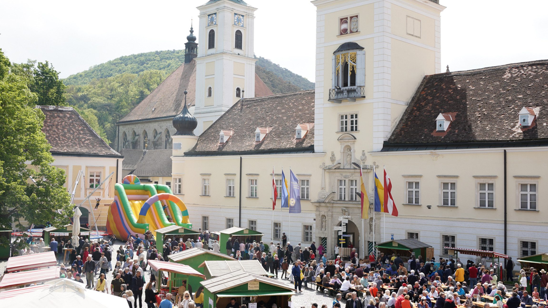 Blick auf das Stift Heiligenkreuz vor dem ein paar Stände und eine Hüpfburg stehen und es stehen und sitzen viele Menschen davor.