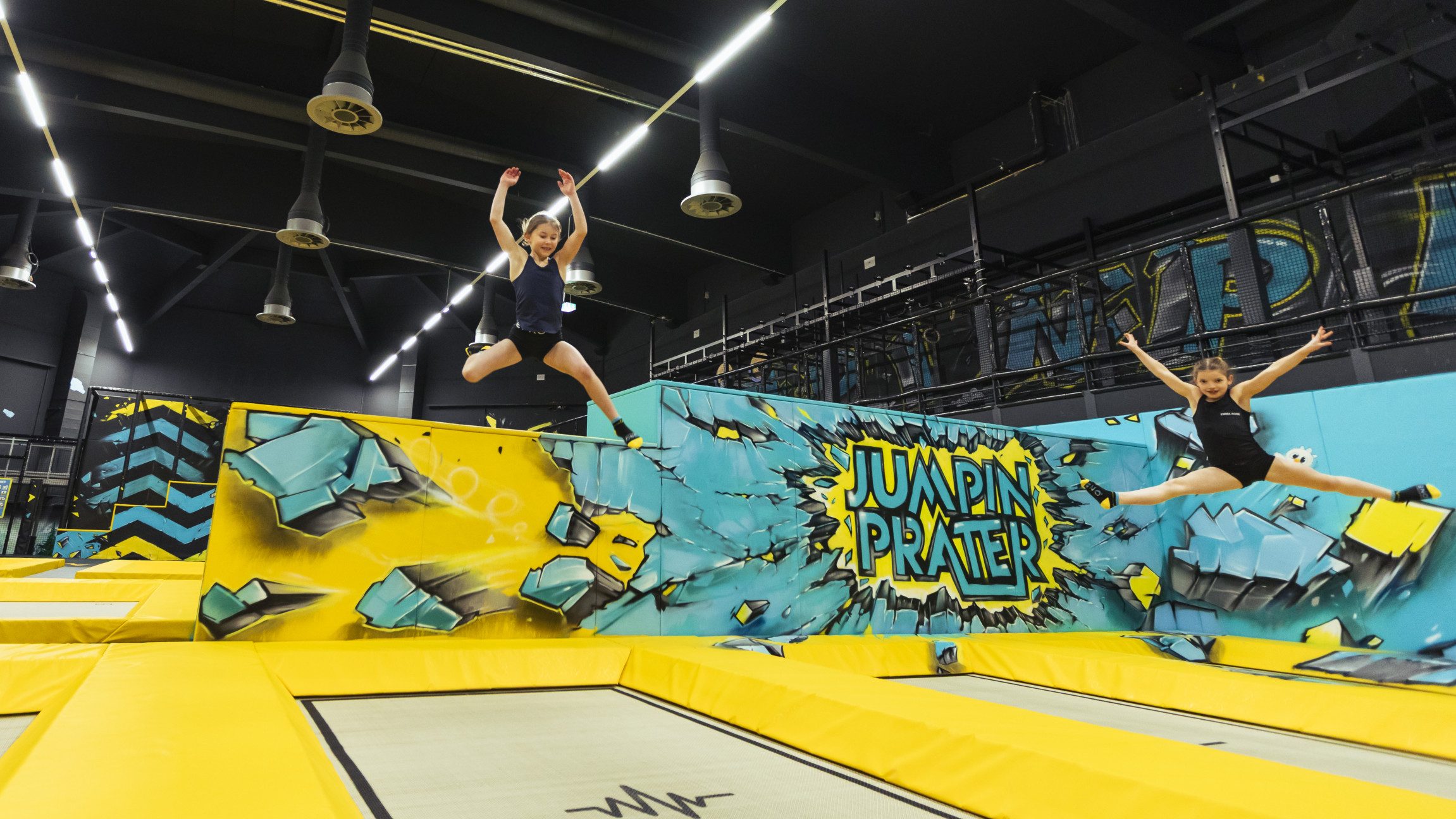 Blick auf zwei Mädchen, die im Jumpin Prater auf zwei Trampolinen hüpfen.