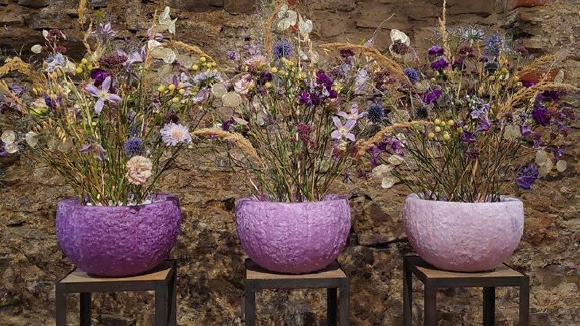 Drei kunstvoll arrangierte Blumensträuße in violetten und rosafarbenen Gefäßen stehen vor einer historischen Steinmauer. Die floralen Werkstücke verbinden zarte Blüten, Gräser und Naturmaterialien zu harmonischen Arrangements.