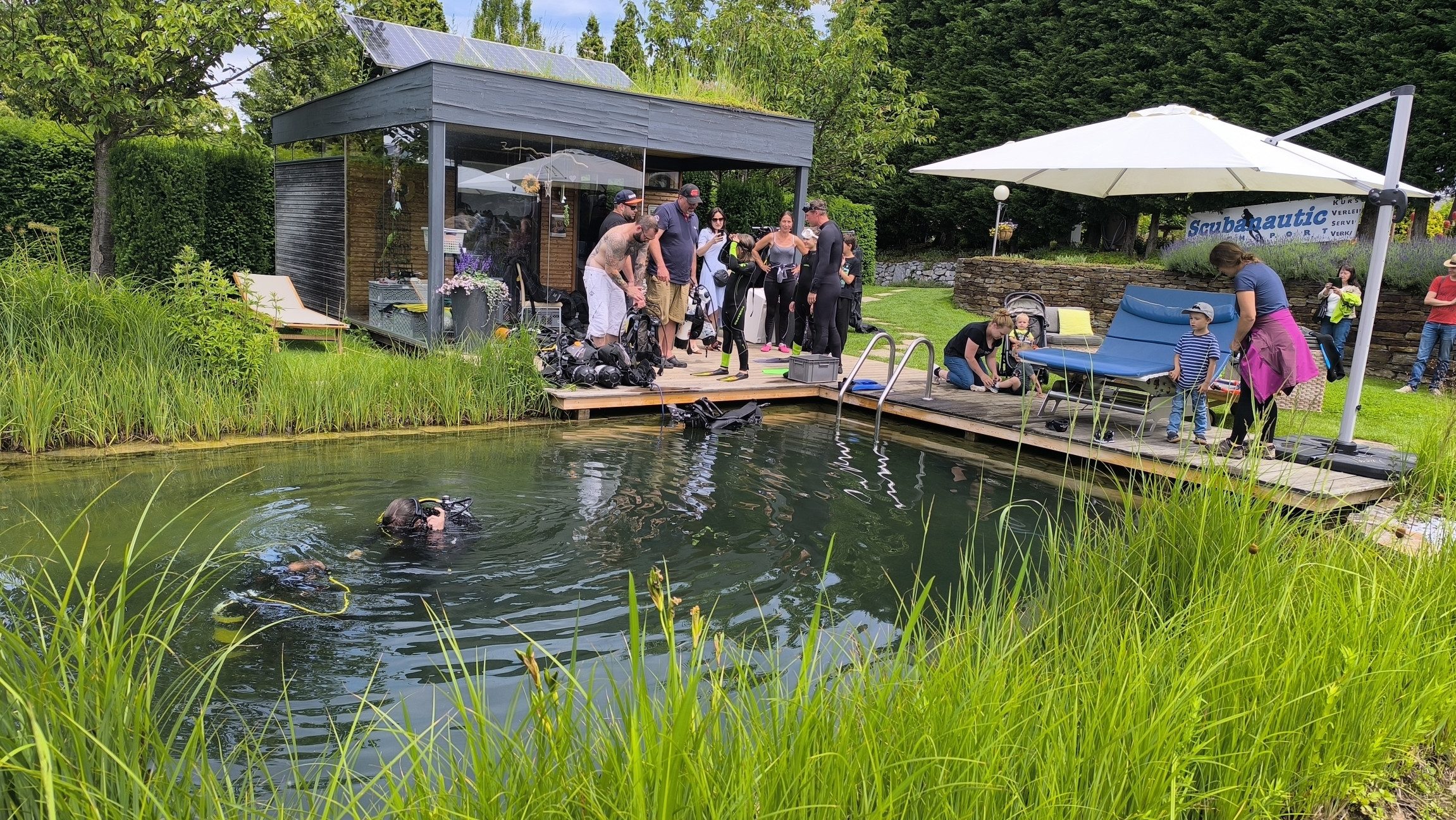 Blick auf einen Naturpool in dem zwei Personen tauchen und mehrere Personen darum herum stehen.