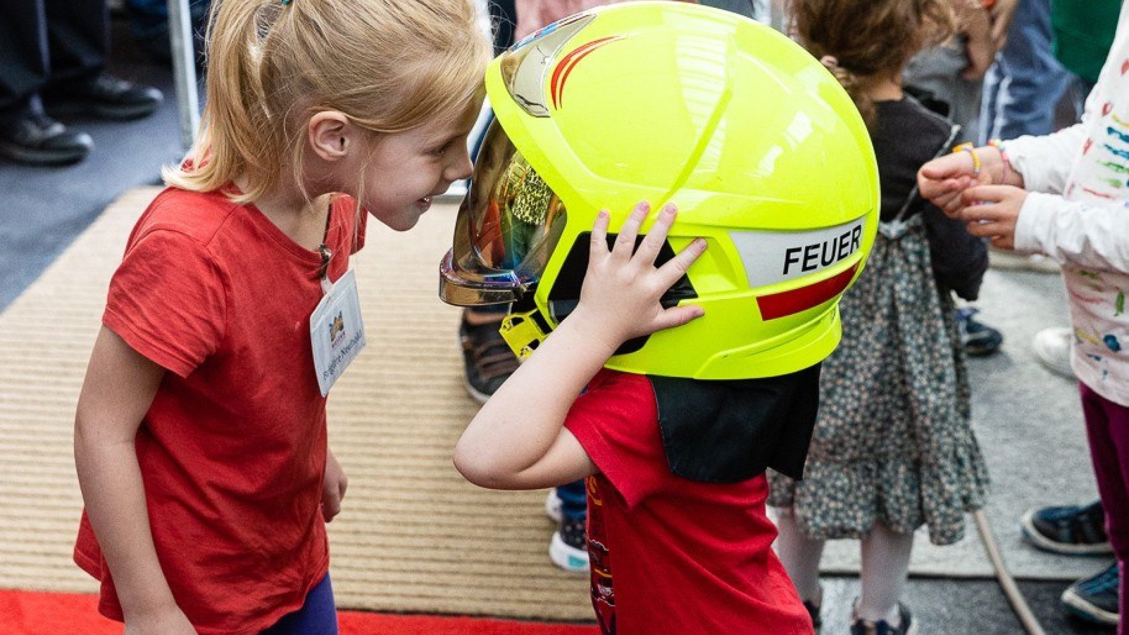 Ein Kind hat einen großen Feuerwehrhelm auf und das andere Kind sieht durch die Schutzbrille des Helms.