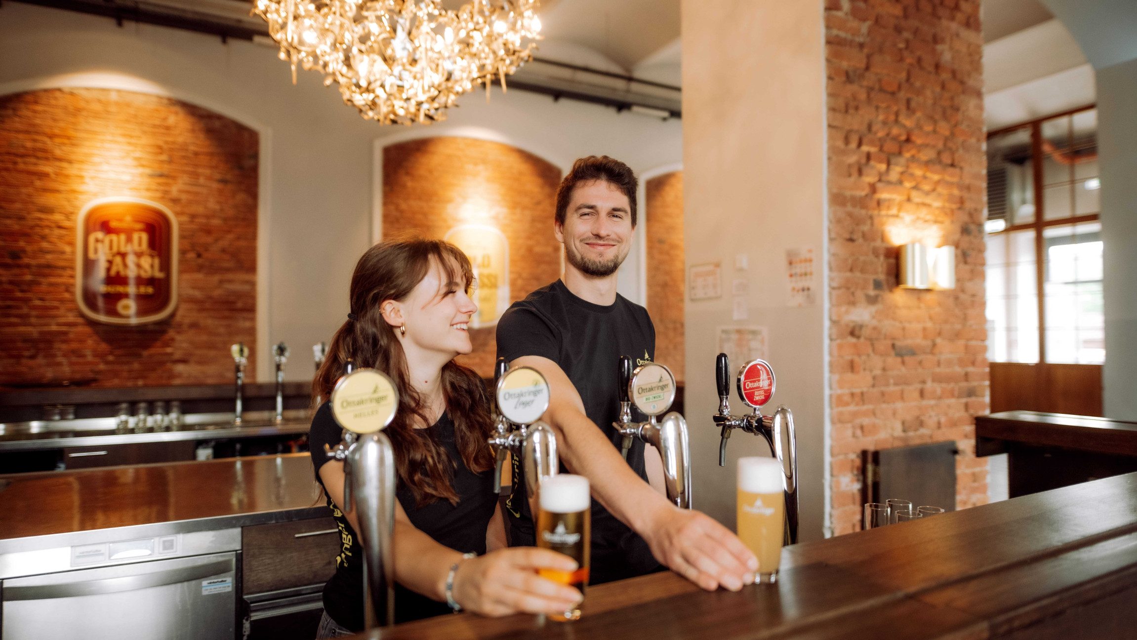 Zwei Personen stehen hinter einer Bar mit einer Zapfsäule und stellen zwei frisch gezapfte Biere auf den Tresen.