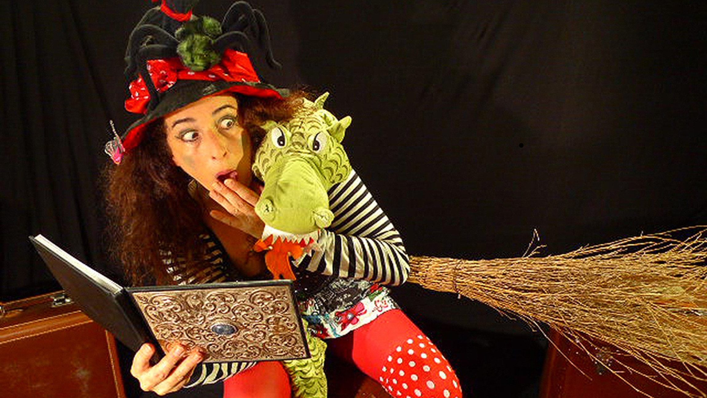 Foto einer als Hexe verkleideten Frau, die ein Buch, einen Stofftier-Drachen und einen Hexenbesen in der Hand hält.