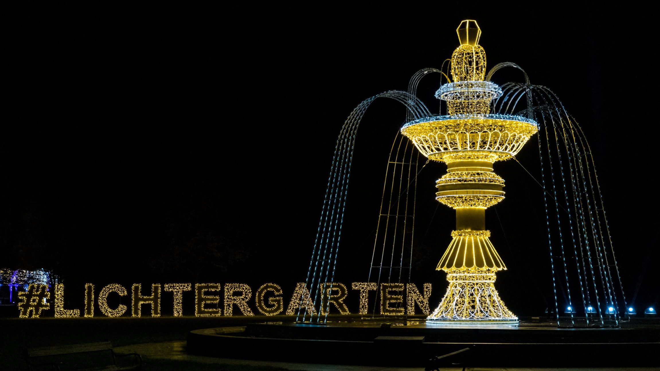 Blick auf einen beleuchteten Springbrunnen mit beleuchteter Schrift "Lichtergarten" im Hintergrund.