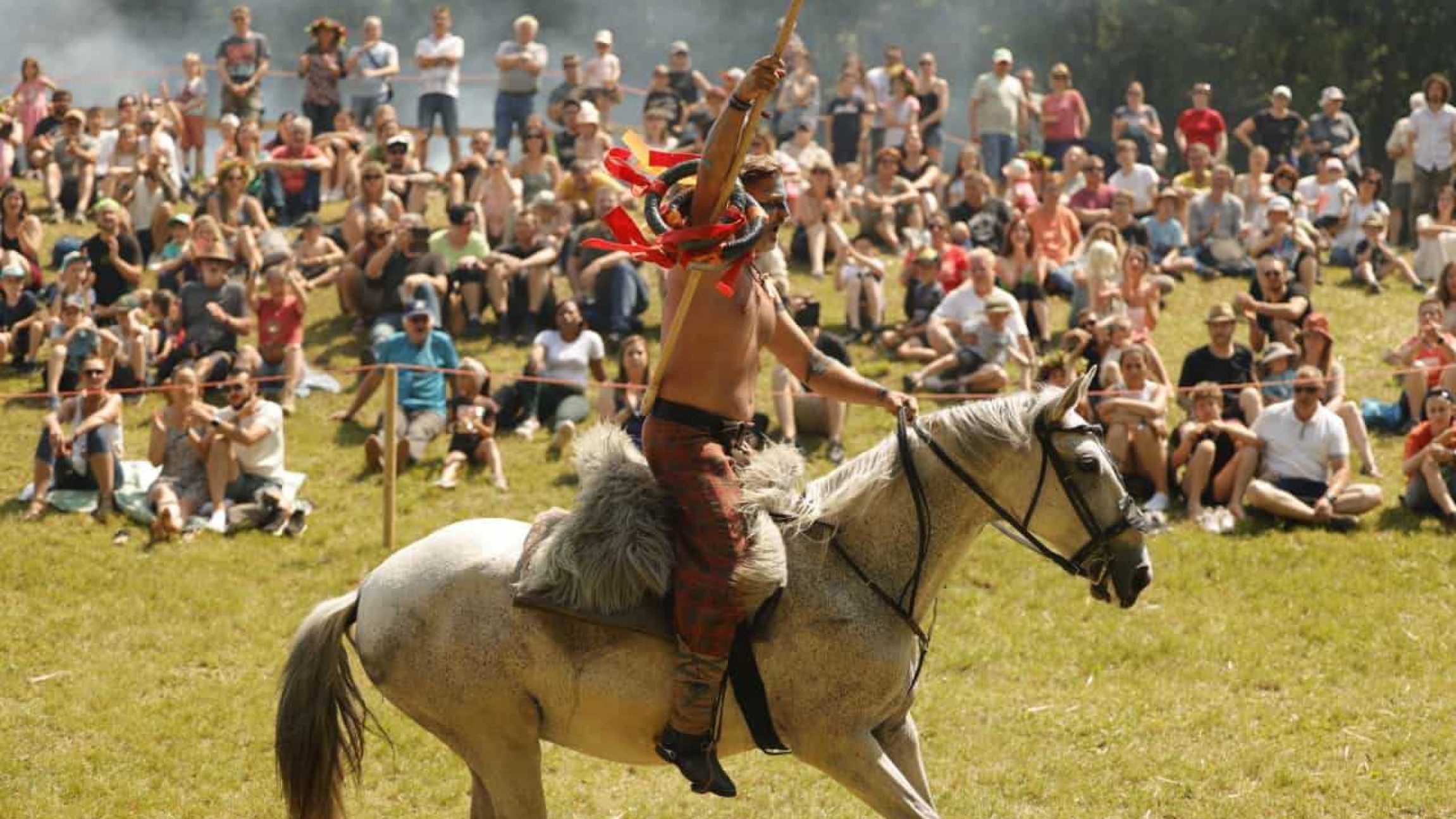 Ein Mann mit nacktem Oberkörper und Kriegsbemalung reitet auf einem Pferd und im Hintergrund sehen viele Personen zu.