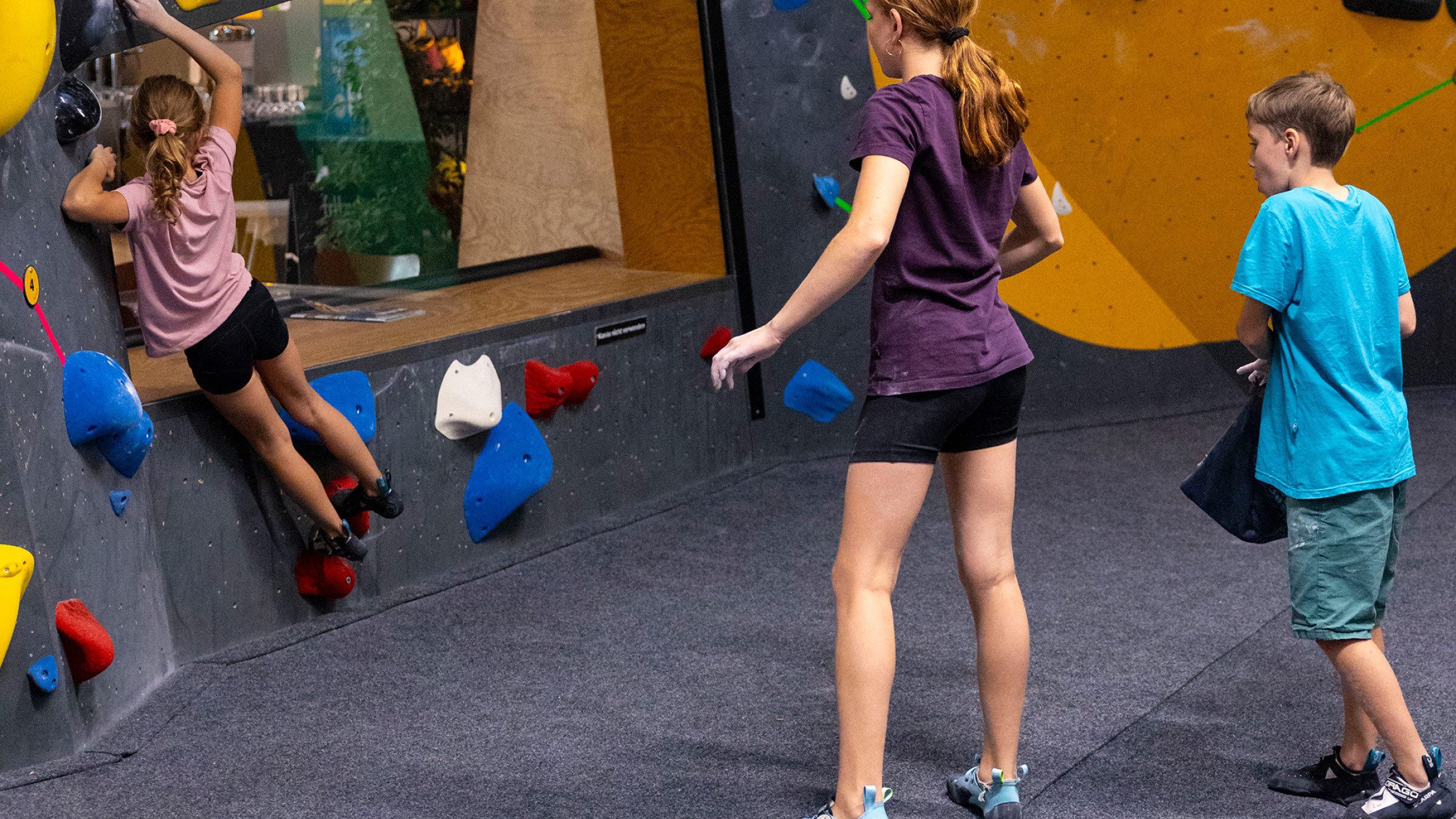 Ein Kind bouldert und zwei weitere Kinder sehen zu.