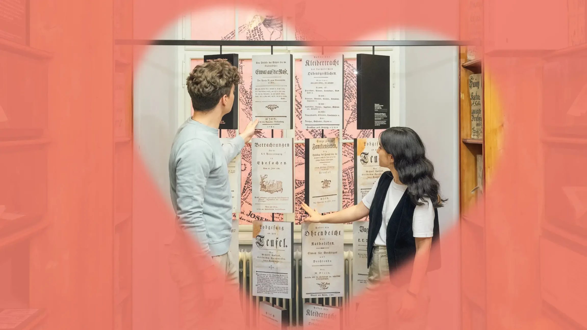 Blick durch ein rosa Herz auf einen Mann und eine Frau, div vor einer Wand mit Urkunden stehen.