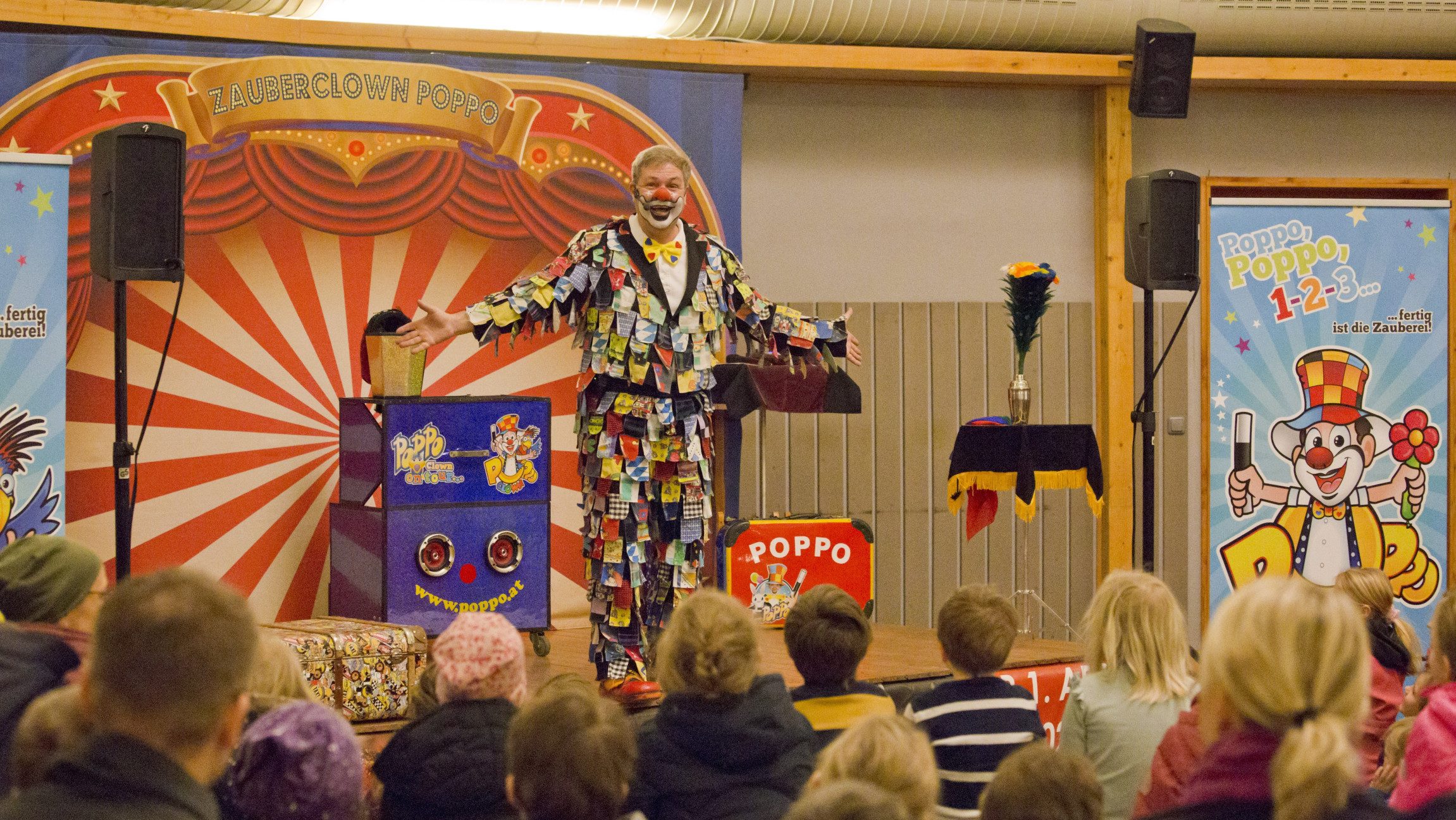 Blick auf einen Clown der auf einer Bühne steht vor der Kinder und Erwachsene sitzen.