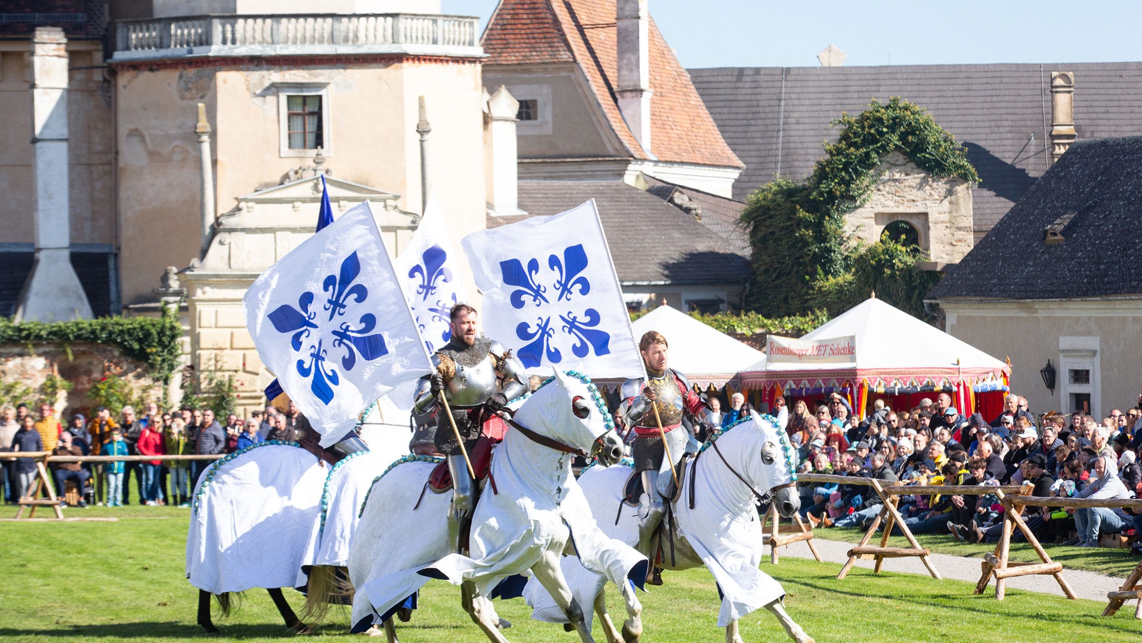 Blick auf ein Rittertournier, bei dem zwei Reiter mit großen Flaggen reiten.