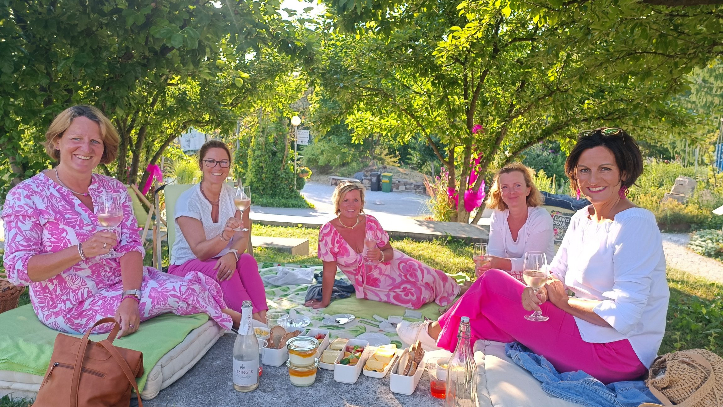 Gruppenfoto von Damen, die mit Rosegläsern auf einer Picknickdecke sitzen und in rosa gekleidet sind.