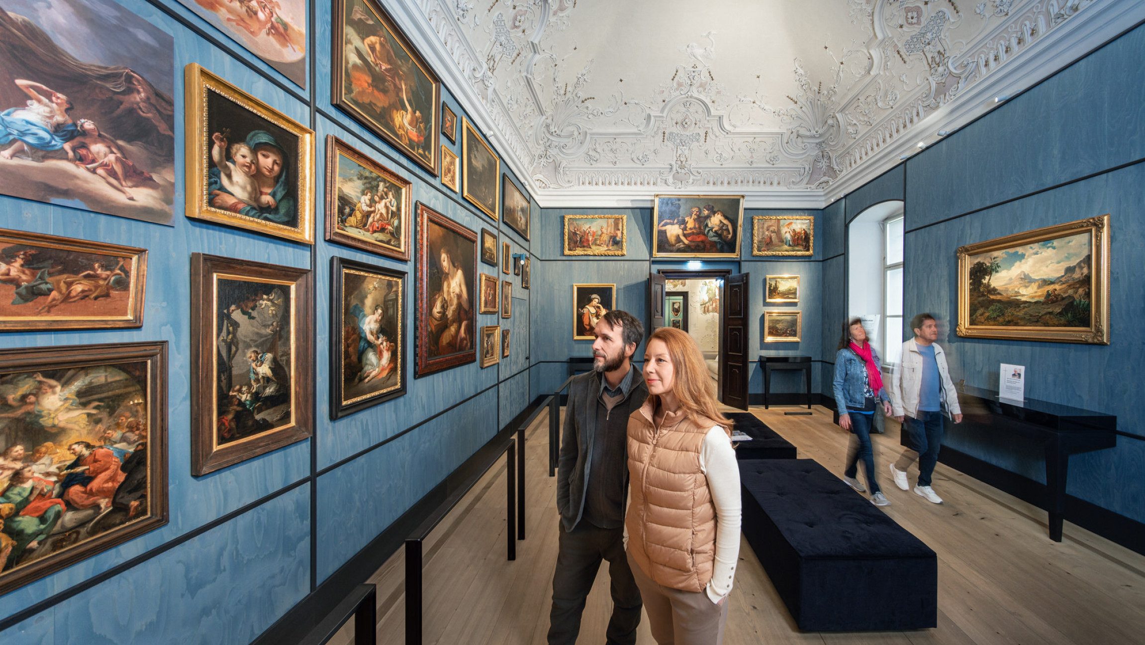 Zwei Erwachsene stehen in einem Ausstellungsraum mit Bildern und sehen sich eines an.