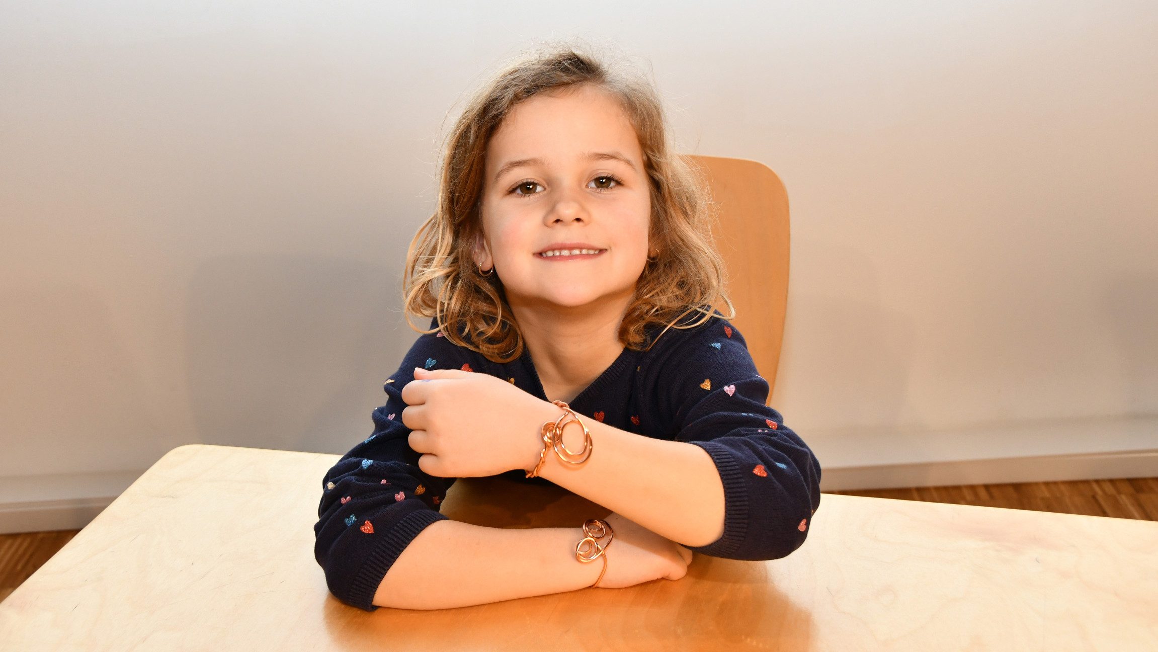 Foto von einem Kind, das an einem Tisch sitzt und Armbänder am Handgelenk herzeigt.