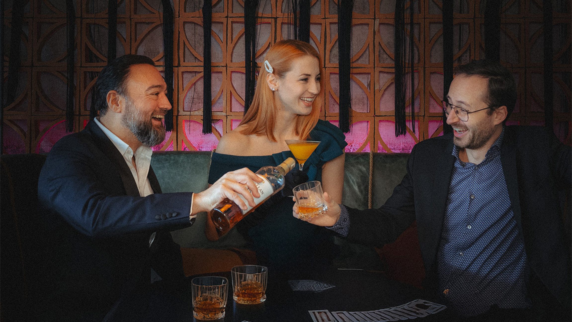 Blick auf zwei Herren und eine Dame, die gerade Cocktails trinken und ein Herr schenkt dem anderen etwas ein.
