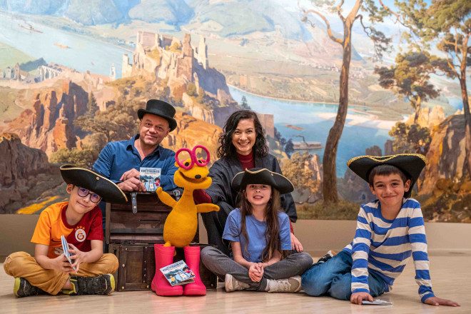 Ein Gruppenfoto von 2 Erwachsenen, 3 Kindern mit Piratenhüten und einem Mascottchen vor einem Hintergrund wo ein Schloss auf einem Berg über einem Fluss zu sehen ist.