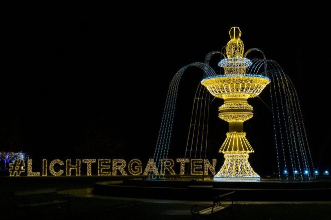 Blick auf einen beleuchteten Springbrunnen mit beleuchteter Schrift "Lichtergarten" im Hintergrund.