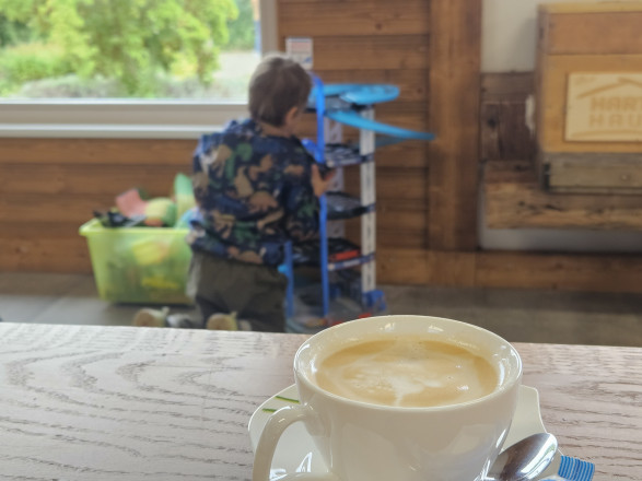 Im Vordergrund des Bildes sieht man eine weiße Tasse mit Kaffee befüllt auf einem Tisch und im Hintergrund sieht man ein Kind mit Spielzeug spielen