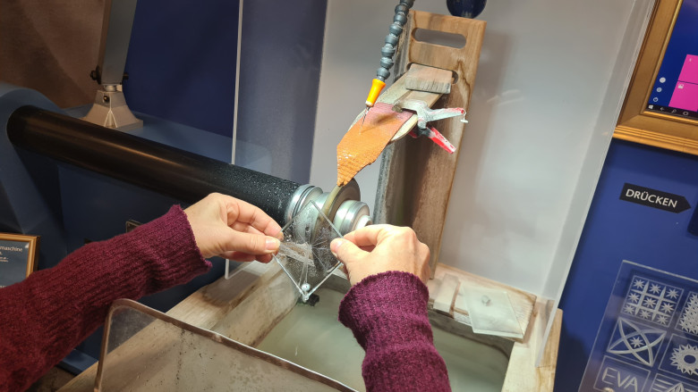 Hände bei der präzisen Glasbearbeitung an einer Schleifmaschine – ein Glasstück wird mit Wasser gekühlt und sorgfältig geschliffen. Traditionelle Handwerkskunst beim Glasschleifen im Kristallium in Niederösterreich, gratis erlebbar mit der Niederösterreich-CARD.