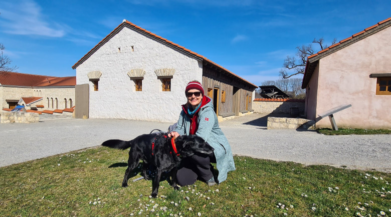 Eine Besucherin kniet mit zwei schwarzen Hunden auf einer Wiese vor rekonstruierten römischen Gebäuden in der Römerstadt Carnuntum. Die historische Anlage ist an einem sonnigen Tag unter blauem Himmel zu sehen.
