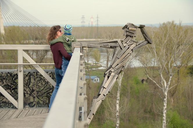 Frau mit Kind auf Plattform, Blick in Natur, abstraktes Holzkunstwerk rechts im Bild