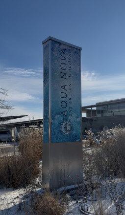 Schild des Aqua Nova Erlebnisbads vor dem modernen Gebäude, umgeben von verschneiten Gräsern unter blauem Himmel.