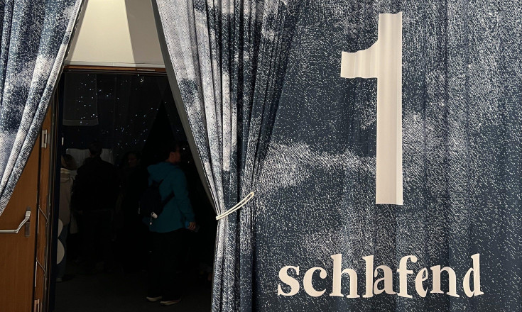 Grauer Vorhang mit dem Schriftzug schlafend, darüber hängt eine Uhr und dahinter befindet sich eine Türe