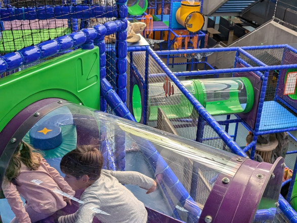 Zwei Kinder spielen in einer Glasröhre des Spielturms, dahinter die Spielwelt