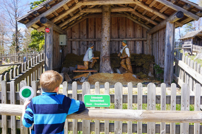 Ein Kind betrachtet eine interaktive Holzstation mit geschnitzten Figuren in einer Erlebniswelt. Die Station zeigt eine Szene aus der traditionellen Holzarbeit in einer Holzhütte.