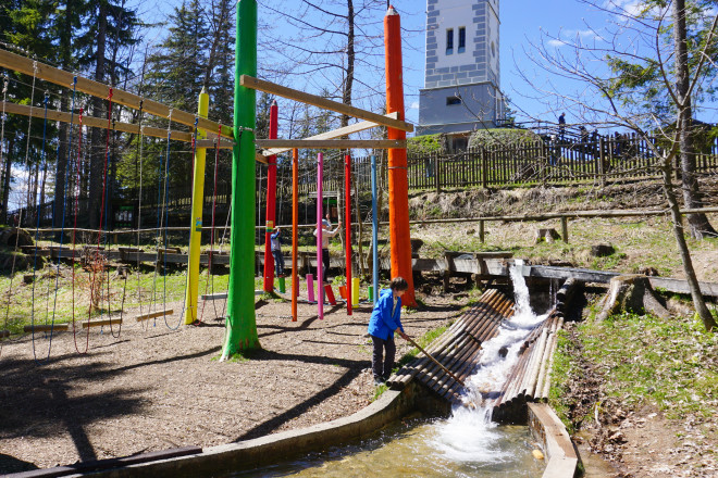 Kinder spielen auf einem Erlebnis-Spielplatz mit bunten Kletterelementen und Wasserspielbereich im Wald. Im Hintergrund steht ein Aussichtsturm unter blauem Himmel.