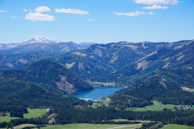 Panoramablick auf eine Berglandschaft mit bewaldeten Hügeln und dem Erlaufsee im Tal. Im Hintergrund sind weitere Berge unter blauem Himmel mit vereinzelten Wolken zu sehen.