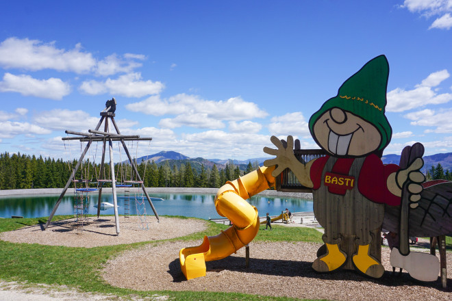 Spielplatz auf einem Berg mit großem Zwergen-Maskottchen, gelber Rutsche und Klettergerüst vor einem kleinen Bergsee. Im Hintergrund sind Wälder und Berggipfel unter blauem Himmel mit weißen Wolken zu sehen.