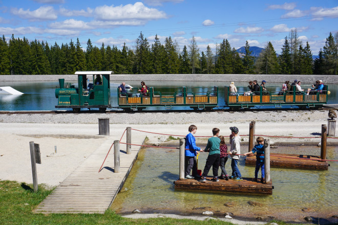 Kinder spielen auf einem Wasserspielplatz mit Holzfloß und Wasserbecken, während im Hintergrund eine kleine Parkeisenbahn an einem Bergsee vorbeifährt. Umgeben ist die Szene von Wald und Berglandschaft.