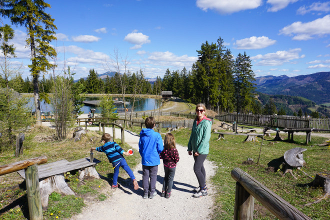Familie mit Kindern spaziert auf einem Weg durch ein Berg-Erlebnisareal mit Blick auf einen kleinen See, Wälder und Berge. Die Landschaft liegt unter blauem Himmel mit vereinzelten Wolken.