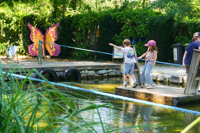 Kinder ziehen sich mit einem Seil auf einem Holzfloß über einen Teich im Kittenberger Erlebnisgärten. Im Hintergrund sorgen bunte Schmetterlingsdekorationen und üppige Pflanzen für eine spielerische Gartenatmosphäre.