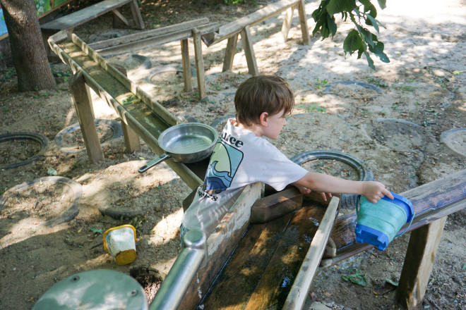 Ein Kind spielt an einer Wasserbahn mit Holzrinnen und gießt Wasser mit einem Eimer in den Wasserspielbereich der Kittenberger Erlebnisgärten. Die Spielstation lädt zum Experimentieren mit Wasser ein.