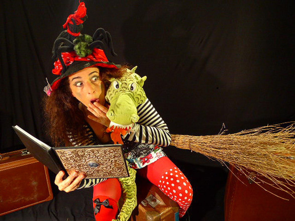 Foto einer als Hexe verkleideten Frau, die ein Buch, einen Stofftier-Drachen und einen Hexenbesen in der Hand hält.