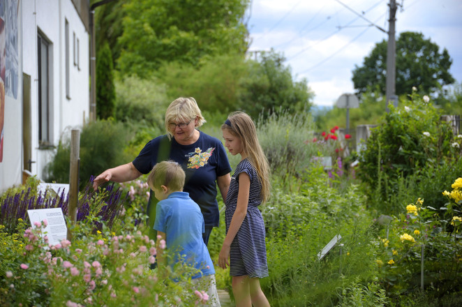Foto einer Dame, die mit zwei Kindern im Kräutergarten steht und ihnen etwas zeigt.