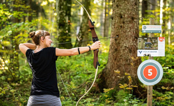 Blick auf eine Dame, die im Wald steht und mit Pfeil und Bogen auf etwas zielt.