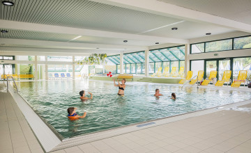 Blick auf ein Schwimmbecken im Hallenbad in dem Kinder mit einem Ball spielen. Darum herum sind ein paar gelbe Liegestühle und viele große Fenster.