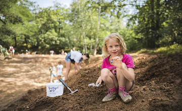 Ein kleines Mädchen sitzt auf einem Erdhaufen mit Grabungsutensilien und hält einen Stein in die Kamera.
