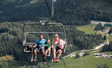 Eine Familie mit einem Kind sitzt auf dem Sessellift und im Hintergrund sieht man das Tal mit Wäldern.