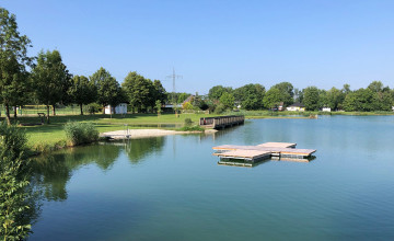 Ein See mit einer Liegewiese und Bäumen im Hintergrund. Auf dem Wasser ist eine kleine Schwimminsel aus Holz.