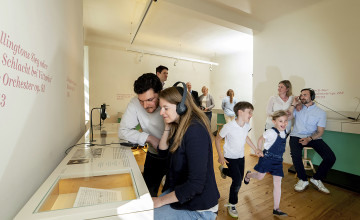 Ausstellungsraum in dem Kinder laufen und zwei Erwachsene mit Kopfhörern etwas anhören.