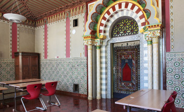 Blick auf einen bunt bemalten Innenraum eines Gebäudes, in dem Klassentische mit roten Stühlen stehen. Eine große Türe mit einem imposanten Türbogen ist zu sehen.