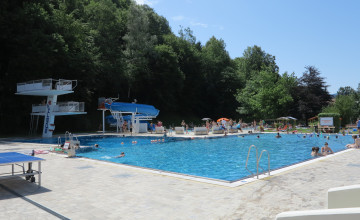 Blick auf ein Schwimmbecken mit Sprungturm, Rutsche und Bäumen im Hintergrund.