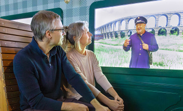 Zwei erwachsene Sitzen in einem Zug auf Holzbänken und statt den Fenstern wird etwas auf einem Bildschirm gezeigt.