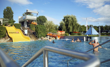 Blick auf einen großen Pool mit einer gelben breiten Rutsche, Stiegen und einer orangen Röhrenrutsche. Personen baden im Wasser.