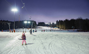 Blicklift auf einen Schlepplift von Hinten mit dem Kinder und Erwachsene bei Flutlichtbetrieb die Piste hinauffahren.